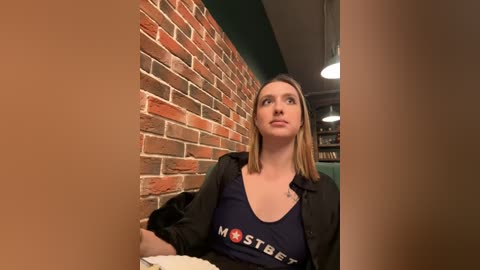 Media: Video of a Caucasian woman with straight, shoulder-length blonde hair, wearing a dark navy blue tank top with \"MostBet\" logo and a black jacket, seated in a brick-walled cafe with modern lighting.