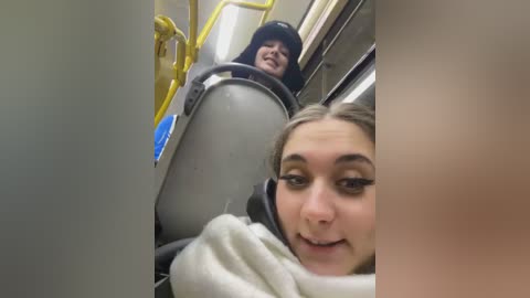 Media: Video of a woman with long brown hair wearing a white sweater, smiling at the camera while riding a subway train with a black helmet. The subway is modern with yellow handrails and a gray seat.