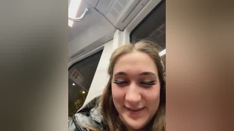 Media: A video of a young woman with long brown hair, wearing black eyeliner and a gray jacket, smiling in a dimly lit subway car.