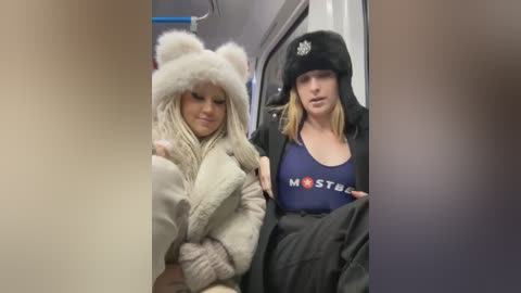 Media: Video of two young women, one wearing a fluffy white bear hat and a beige coat, and the other in a black fur hat and a blue \"Mister\" t-shirt, seated in a subway car.