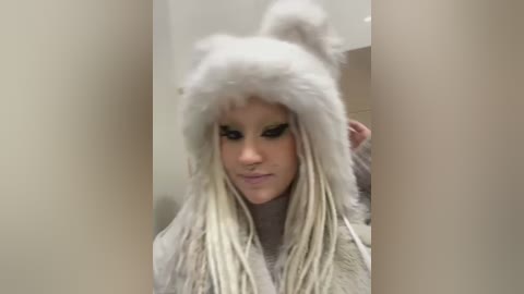 Media: Video of a woman with light skin and long blonde dreadlocks, wearing a fluffy white rabbit hat, standing in a beige-tiled room, looking down, with slightly blurred background.