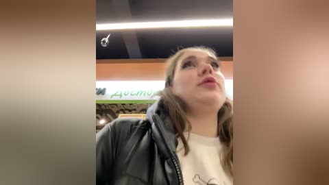 Media: Video of a young woman with light skin, wearing a black leather jacket over a white shirt, looking upward with a neutral expression. Background features a blurred sign and ceiling lights.