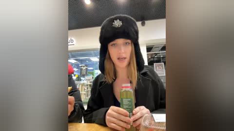 Media: Video of a young woman with light skin and shoulder-length blonde hair, wearing a black fur hat with a red star and a black hoodie, holding a green juice bottle at a grocery store.