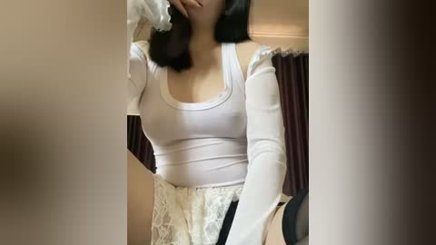 Media: Video of an Asian woman with medium skin tone, wearing a tight white long-sleeve top that reveals her small breasts, lace skirt, and black hair. She is indoors near a dark curtain.