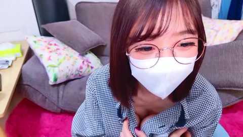 Media: Video of a young Asian woman with straight, shoulder-length brown hair, wearing glasses, a white face mask, and a blue checkered shirt, sitting on a pink rug in a living room with gray furniture and colorful cushions.