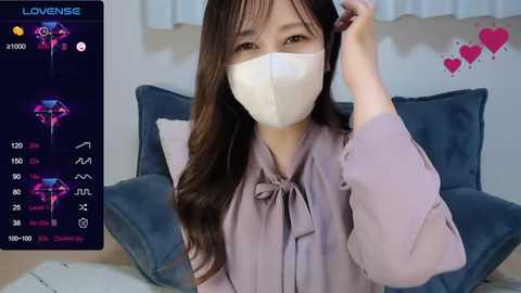 Media: A video of an East Asian woman with long brown hair and a white face mask, wearing a lavender blouse, sitting on a blue couch. A digital overlay shows heart symbols and a glowing device.