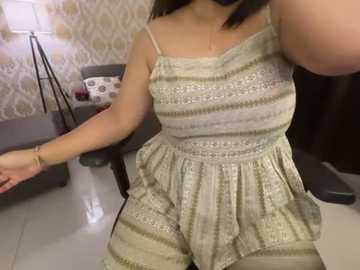 Media: Video of a light-skinned woman with shoulder-length dark hair, wearing a sleeveless, patterned, light green romper, standing in a room with patterned wallpaper, a gray chair, and a black floor lamp.