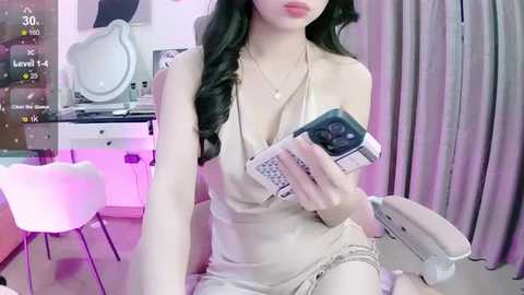 Media: Video of a young woman with long black hair, fair skin, wearing a beige dress, holding a phone, sitting on a chair in a pink-lit room with a white chair and a vanity table.