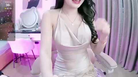 Media: A video of a smiling woman with long, wavy black hair, wearing a revealing, white, halterneck dress, sitting on a modern white chair. The room has a modern decor with a white vanity and pink lighting.