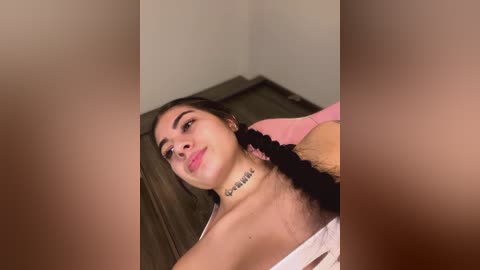 Media: Video of a young, light-skinned woman with long dark hair, wearing a pink cardigan, lying on a wooden floor, partially topless, with a tattoo on her neck.