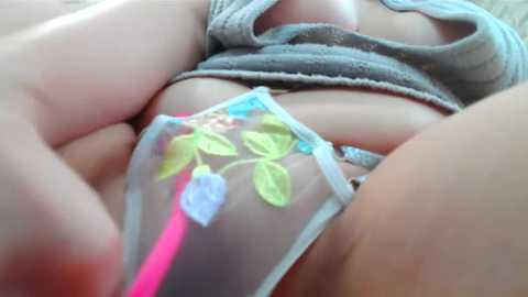 Media: Video of a woman's torso in a sheer, floral-patterned pink bralette with blue leaves and a light grey tank top. Her skin is fair, and her breasts are partially visible through the bra.