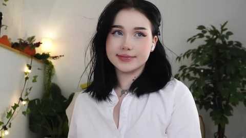 Media: Video of a young woman with fair skin, blue eyes, and shoulder-length black hair, wearing a white button-down shirt. She stands indoors with green plants and string lights in the background.