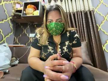 Media: Video of a woman with light brown skin, wearing a green face mask, black floral shirt, and black pants, seated on a brown couch, surrounded by decorative pillows and yellow-patterned wall.