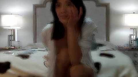 Media: Video of an Asian woman with long black hair, partially nude in a white shirt, sitting on a bed with a cow-print blanket, in a dimly lit bedroom with two bedside lamps.