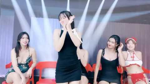 Media: Video of four East Asian women, three in revealing outfits, one in a black dress, seated on red chairs, under white spotlights, smiling.