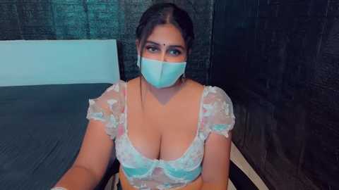 Media: Video of a woman with medium skin tone, wearing a white surgical mask, pastel floral lingerie, and a blue surgical gown, sitting in a dark, brick-walled room.