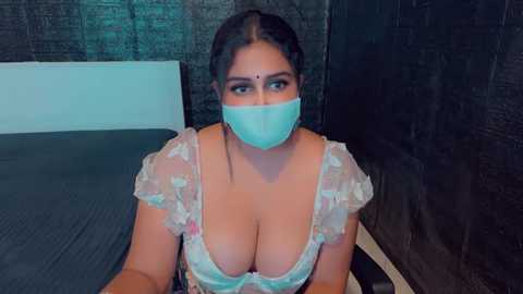 Media: A video of a South Asian woman with medium skin tone, dark hair, and a large bust, wearing a light blue floral top and face mask, sitting on a bed with a black brick wall in the background.