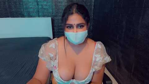 Media: Video of a South Asian woman with dark hair and light skin, wearing a white floral-patterned bra that accentuates her large breasts, a blue surgical mask, and a headband, seated on a bed with a dark headboard and textured wall.