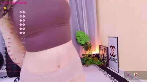 Media: A video of a fair-skinned woman wearing a tight brown crop top and white lace panties, standing in a room with a monstera plant, Chanel logo, and a black and white patterned tablecloth.