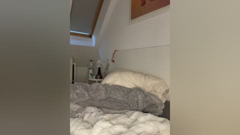 Media: Video of a messy bedroom with a gray comforter, white pillows, a white wall, and a small shelf holding a few items.