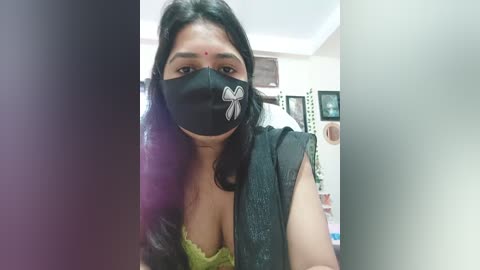 Media: Video of a South Asian woman with medium skin tone, long black hair, wearing a black face mask with a butterfly design, a green lace bra, and a black vest, standing indoors with blurred background.