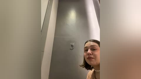 Media: A video of a young woman with light skin and shoulder-length brown hair, wearing a beige sweater, peering through a narrow gap in a white bathroom door. The room has a minimalist, modern design.