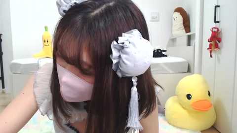 Video of an Asian woman in a maid costume with a pink face mask, sitting on a colorful rug in a white room with plush toys and a stuffed yellow duck.