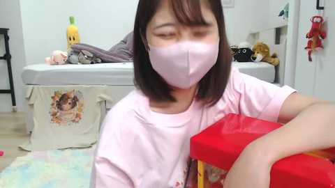 Video of a young Asian girl with shoulder-length black hair, wearing a pink mask, pink t-shirt, and sitting on a soft rug. She is opening a red gift box with a red ribbon in a cozy, cluttered bedroom.