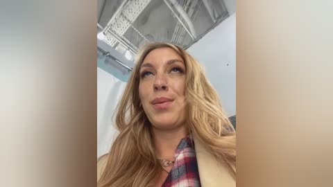 Media: Video of a blonde woman with long, wavy hair, wearing a beige jacket and a plaid shirt, standing in an industrial room with exposed beams and white walls.