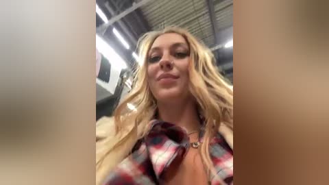 Media: Video of a blonde woman with long, wavy hair, wearing a red and white plaid shirt, smiling confidently indoors with a modern industrial ceiling.