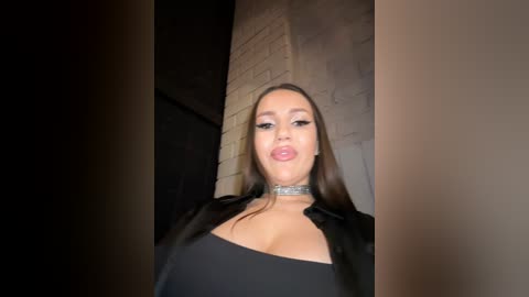 Media: Video of a woman with long dark hair, wearing a black top, silver choker, and black jacket, standing against a dimly lit, textured brick wall.