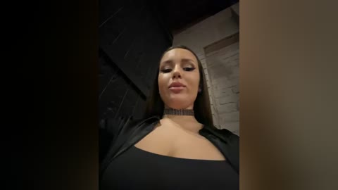 Media: Video of a light-skinned woman with long brown hair, wearing a black choker and low-cut black top, indoors with dim lighting, highlighting her cleavage and confident expression.