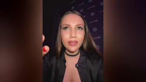 Media: Video of a young, attractive woman with long, straight brown hair, wearing a black blazer and choker, with red nails, against a dark backdrop.