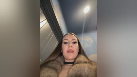 Media: Video of a young woman with long brown hair, wearing a black choker and fur coat, looking up, in a dimly lit room with a blue wall and a bright ceiling light.