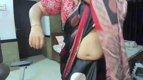 Media: Video of a South Asian woman in a red and black saree, lifting her sari to reveal her belly, in a cluttered room with a table, chair, and electrical outlet.