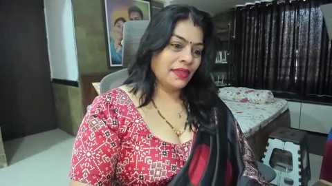 Media: Video of a middle-aged woman with long black hair, wearing a red patterned blouse, sitting in a modern bedroom with a bed, dark curtains, and a framed photo on the wall.