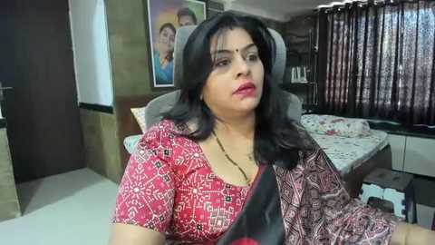 Media: Video of an Indian woman with long black hair, wearing a red and black patterned sari, sitting on a gray chair in a modest, traditionally decorated bedroom with a framed photo on the wall.