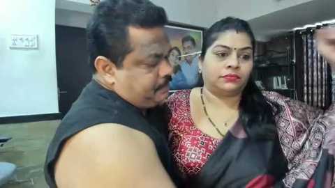 Media: Video of a man in a black tank top and a woman in a red-patterned saree, both with dark hair, in a room with a framed picture on the wall.