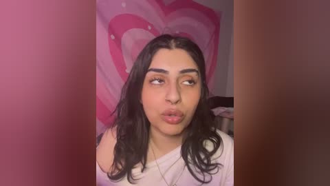 Media: Video of a young woman with medium brown skin, long dark hair, and full lips, pouting against a pink heart backdrop. She wears a white off-shoulder top.