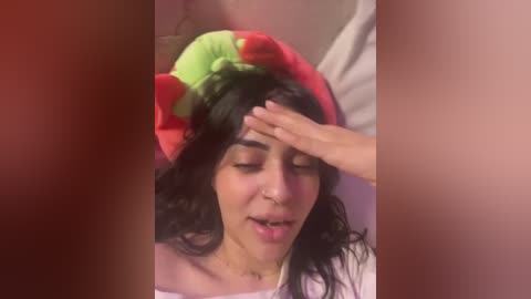 Media: A video shows a young woman with olive skin, wearing a red and green clown hat, lying on a bed with her eyes closed and hand on her forehead, suggesting fatigue or illness.