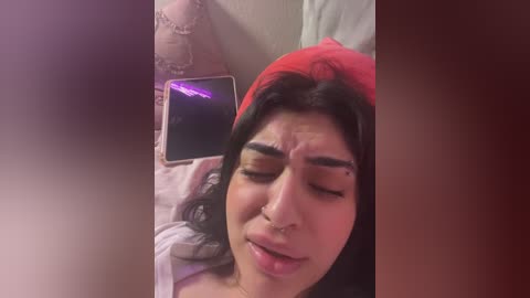 Media: Video of a young woman with medium skin tone, long black hair, wearing a red beanie and a white top, lying on a bed, phone in hand, with a slightly annoyed expression.