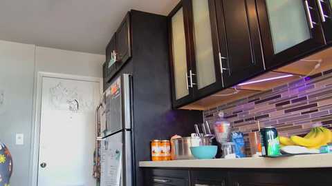 Media: Video of a modern kitchen with dark wooden cabinets, stainless steel appliances, and a mosaic tile backsplash. Countertop cluttered with a bowl, bananas, and utensils.