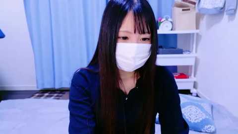 Media: A video of an Asian woman with long black hair and a white surgical mask, wearing a navy-blue blouse, sitting indoors with a white wall, blue curtains, and a white shelf in the background.