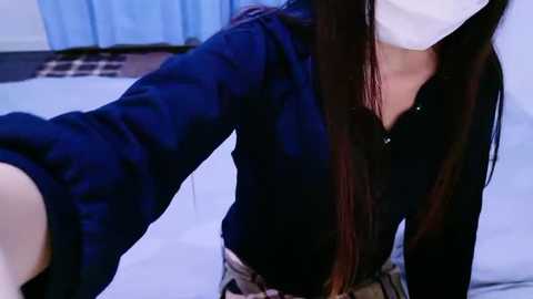 Media: Video of a young woman with long dark hair, wearing a blue shirt, white mask, and plaid skirt, taking a selfie in a bright room with blue curtains.