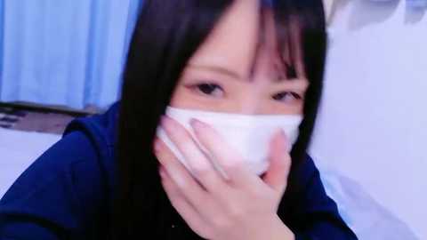 Media: Video of an East Asian woman with straight black hair, wearing a white surgical mask, blue coat, and blue curtains in the background. She is holding her face with her left hand.