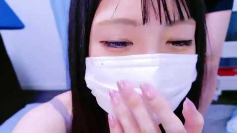 Media: A close-up video of an Asian woman with long black hair, wearing a white surgical mask, blue eyeshadow, and a black top, with a blurred background.