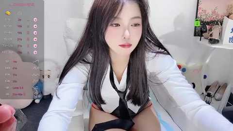 Media: Video of an East Asian woman with long black hair, wearing a white blouse and black tie, in a messy room with a calendar, shoes, and toys.