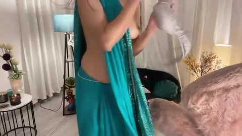 Media: Video of a woman with light skin, wearing a teal sari, partially lifting it to reveal her bare back, in a modern bedroom with beige curtains, a white lamp, and a bed.