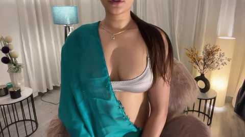 Media: Video of a young woman with light skin, wearing a teal wrap and a white bra, sitting in a beige chair, surrounded by modern decor in a softly lit room.