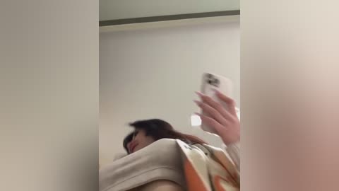 Media: Video of a woman with long dark hair, wearing a beige sweater with orange and yellow patterns, taking a selfie in a dimly lit room with off-white walls.